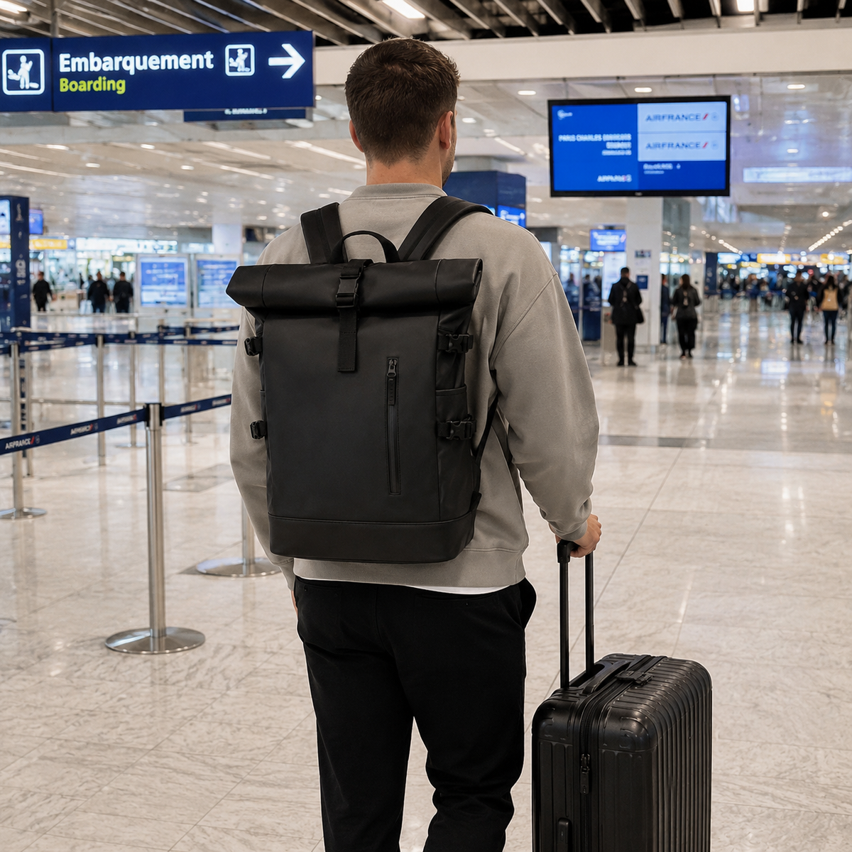 Voyageur de dos dans un hall d'aéroport, sac à dos roll-top noir sur le dos, valise noire à la main